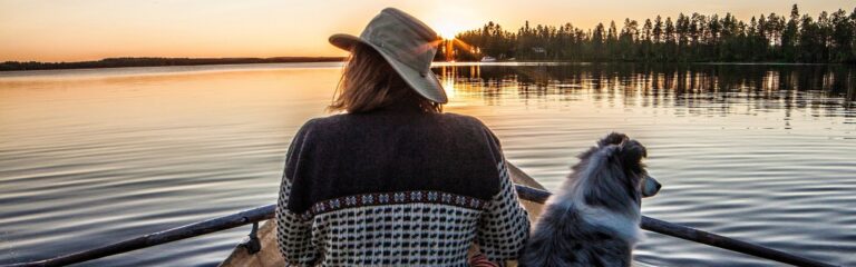 A man and a dog on a rowboat on a still lake at sunset.