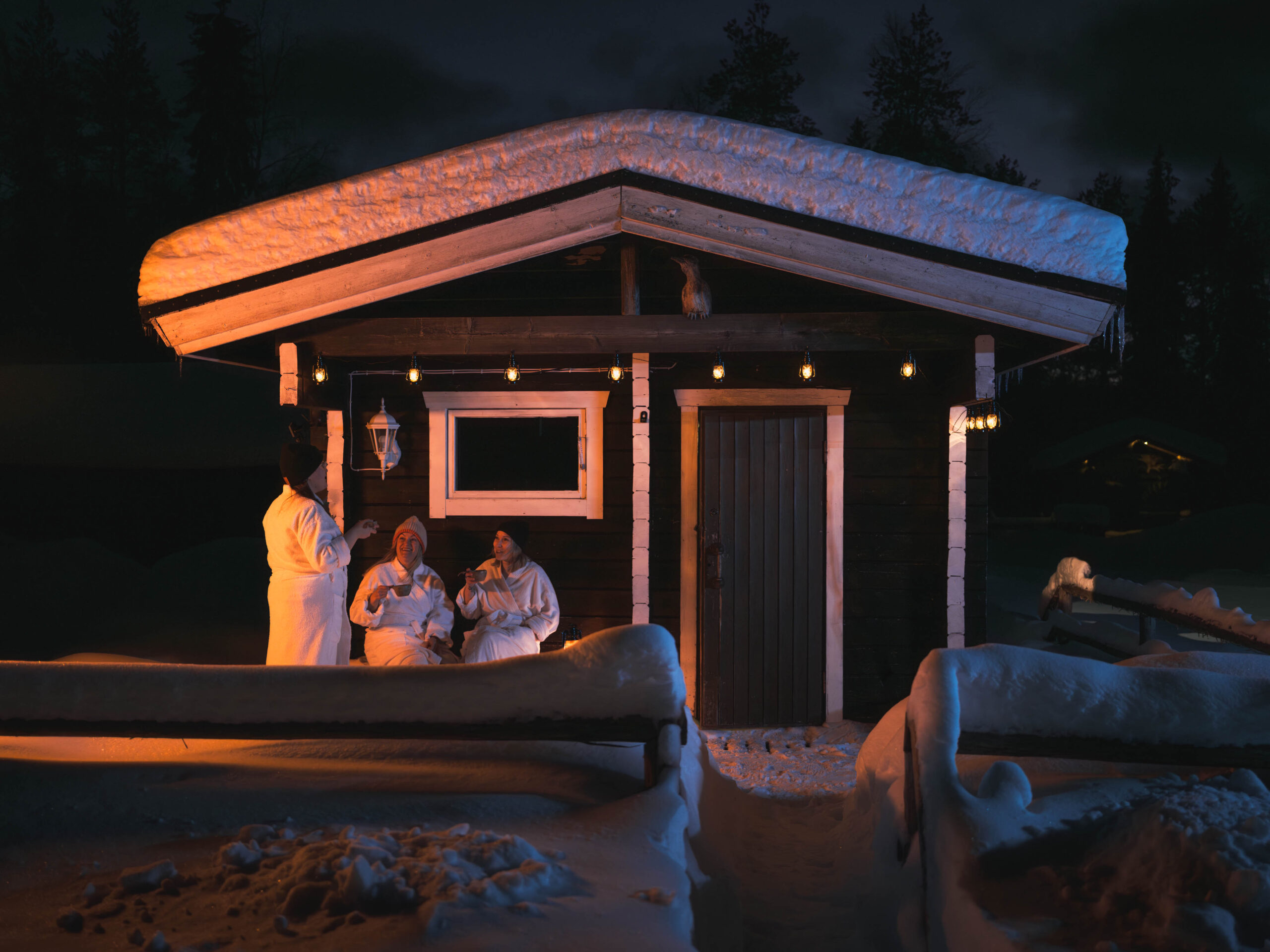 Women enjoying fresh air in robes in front of a traditional Finnish sauna building.
